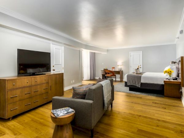 A spacious hotel room with a bed, TV on a dresser, sofa, small table, desk with chair, and wooden flooring. The room has soft lighting and minimal decor.