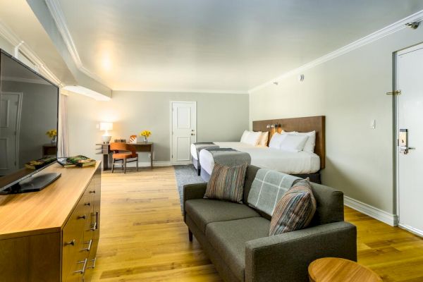 A modern hotel room features a large bed, a sofa, a wooden dresser with a TV, a desk with a chair, and a door.