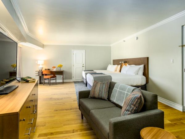 A modern hotel room features a large bed, a sofa, a wooden dresser with a TV, a desk with a chair, and a door.