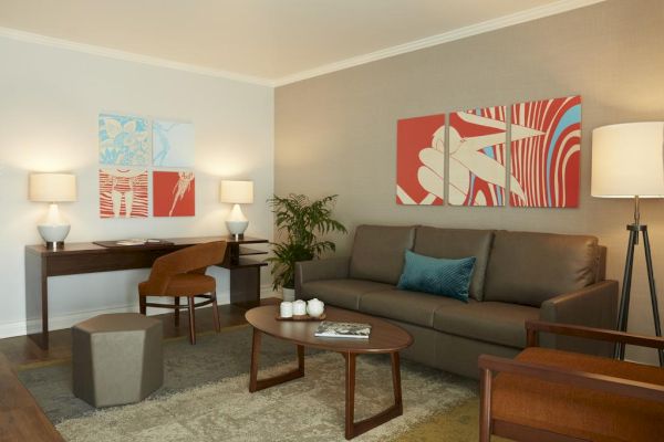 A modern living room with a sofa, coffee table, desk, chairs, lamps, abstract wall art, and a potted plant.