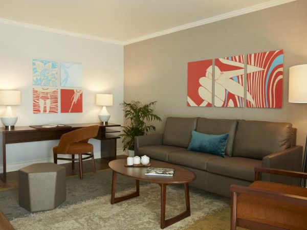 A modern living room with a sofa, coffee table, desk, chairs, lamps, abstract wall art, and a potted plant.