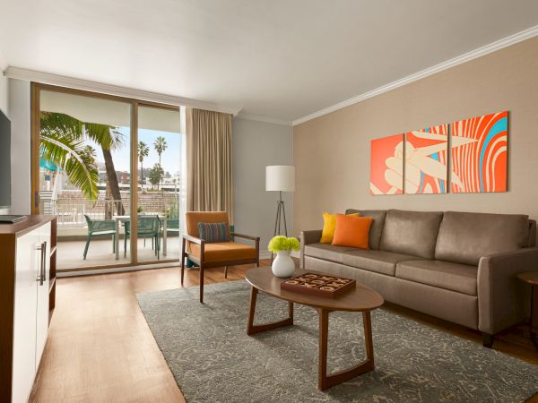 A modern living room with a beige sofa, colorful wall art, a wooden coffee table, a gray rug, and a sliding glass door opening to a balcony.