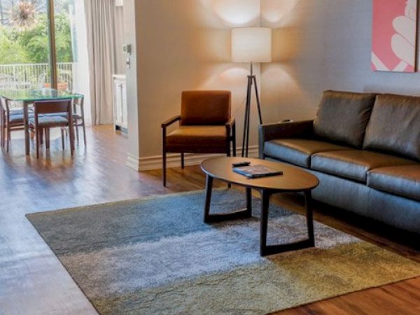 A modern living room with a sofa, armchair, coffee table, floor lamp, and a rug. A dining area with a table and chairs is seen in the background.