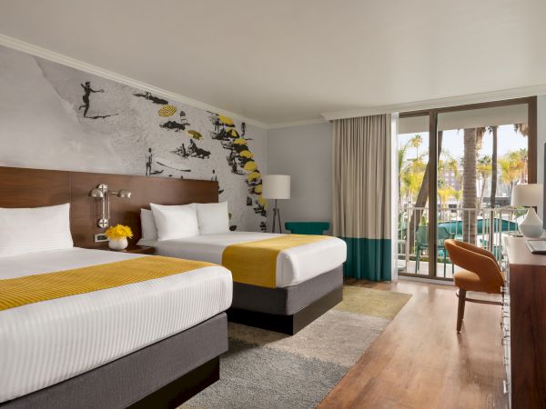 A modern hotel room with two double beds, a desk, a colorful mural above the beds, a balcony view, and warm lighting.