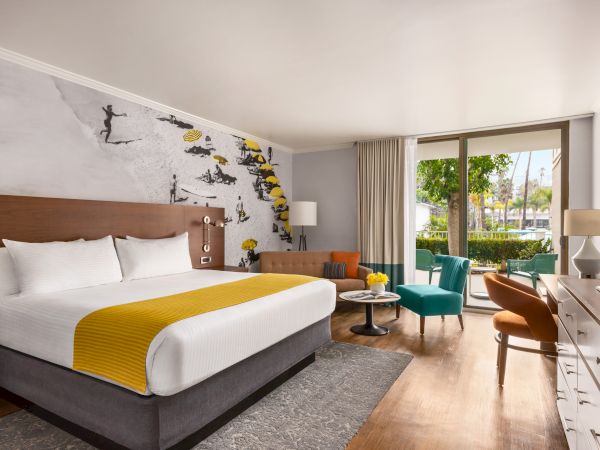 A stylish hotel room with a king bed, mustard-yellow throw, colorful seating, a desk, and a mural wall leading to a balcony.