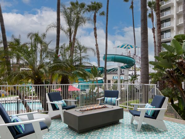A sunny hotel pool deck with lounge chairs, a fire pit, palm trees, and a distant water slide beside the buildings.