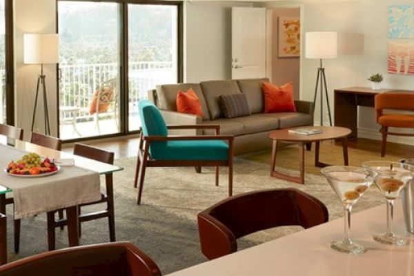 A modern living room with a gray sofa, orange and blue chairs, dining table, lamps, and a bar counter with martini glasses, overlooking a balcony.