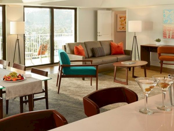 A modern living room with a gray sofa, orange and blue chairs, dining table, lamps, and a bar counter with martini glasses, overlooking a balcony.