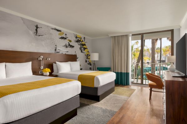 This image shows a hotel room with two beds, yellow accents, a surf-themed wall mural, and a balcony with a view of palm trees.