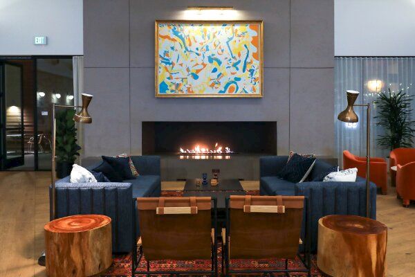 A modern living room with a fireplace, abstract painting, blue sofas, wooden chairs, and round wooden tables. The room also has floor lamps and plants.