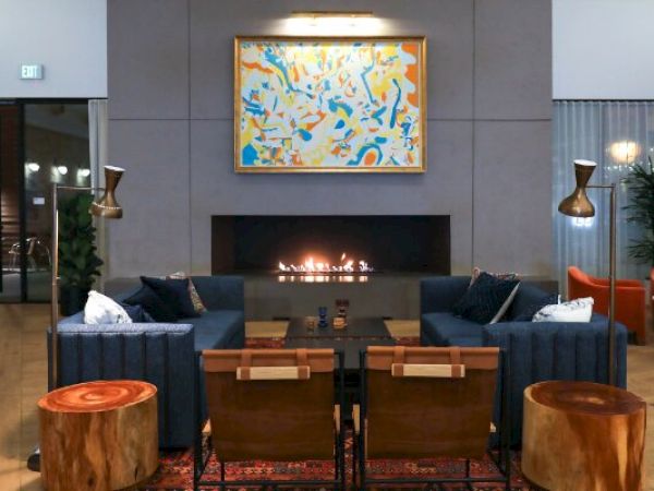 A modern living room with a fireplace, abstract painting, blue sofas, wooden chairs, and round wooden tables. The room also has floor lamps and plants.