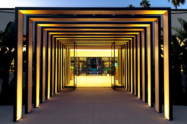 A well-lit geometric passageway made of metal frames leads to a building's entrance. The structure creates a visually appealing, tunnel-like effect.