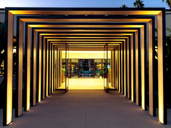 A well-lit geometric passageway made of metal frames leads to a building's entrance. The structure creates a visually appealing, tunnel-like effect.