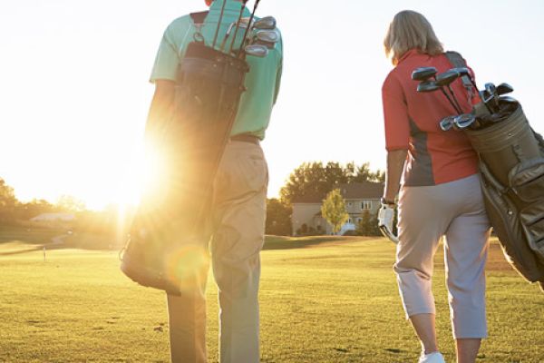 Two individuals are walking on a golf course carrying golf bags, with the sun shining brightly behind them.