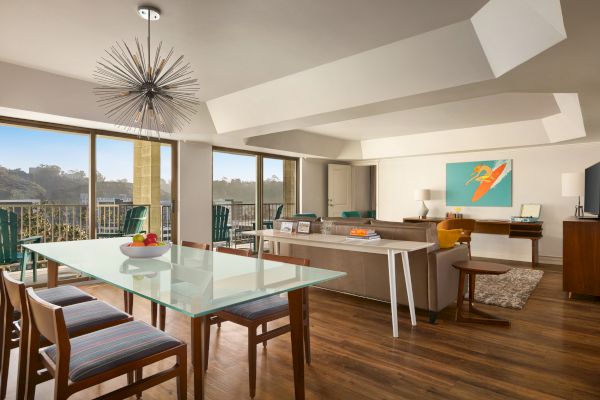 A modern living room with a glass dining table, stylish chairs, and a large sofa. There's a vibrant artwork on the wall and a unique chandelier.
