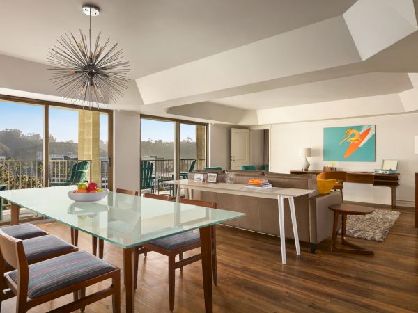 A modern living room with a glass dining table, stylish chairs, and a large sofa. There's a vibrant artwork on the wall and a unique chandelier.