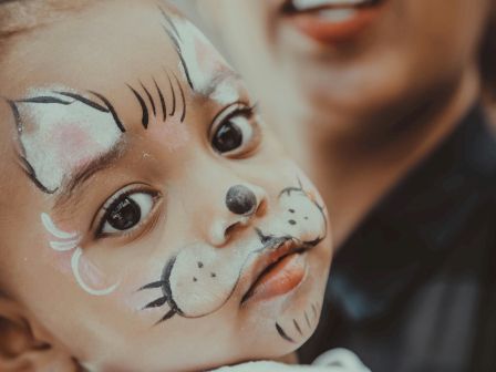 A cute child with face paint resembling a cat: whiskers, nose, and ears drawn around the cheeks and nose, held by a smiling adult in the background.