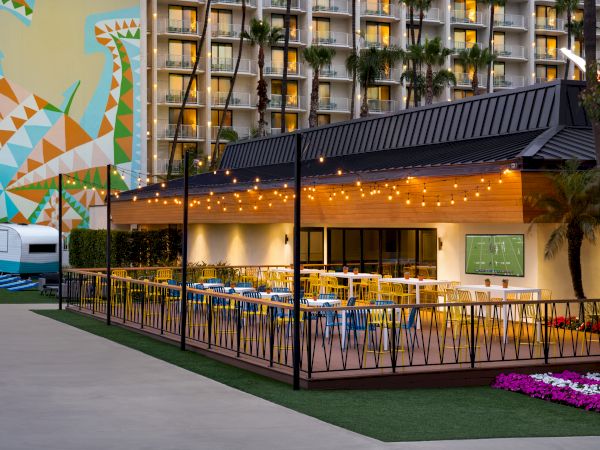 An outdoor restaurant with string lights and colorful seating, adjacent to a hotel with balconies and a large geometric mural on a building side.