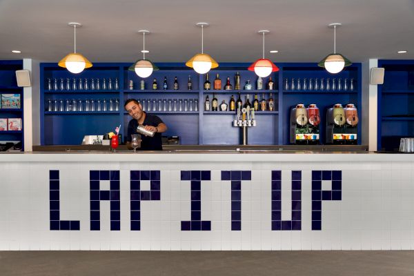 A bartender is preparing a drink behind a counter with the phrase "LAP IT UP" in large letters; bottles and drink dispensers are displayed on shelves.