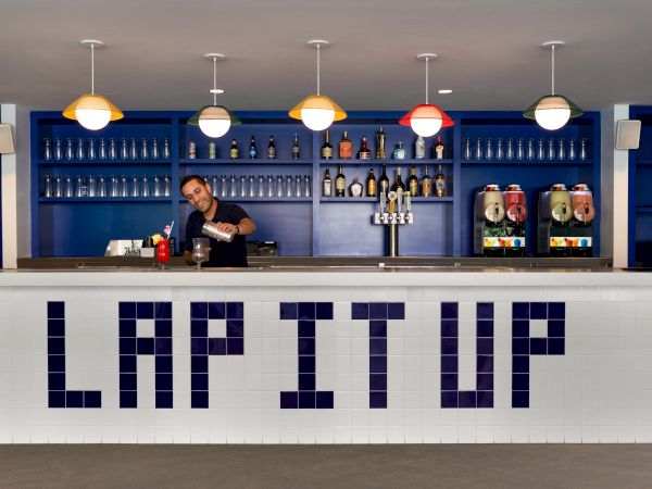 A bartender is preparing a drink behind a counter with the phrase "LAP IT UP" in large letters; bottles and drink dispensers are displayed on shelves.