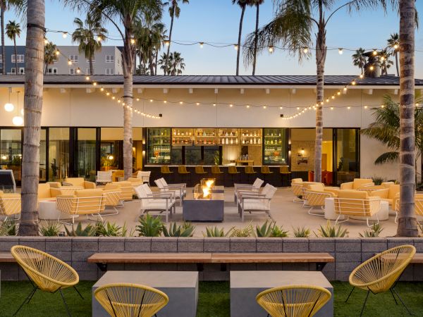 An outdoor seating area with yellow chairs, string lights, and palm trees. There's a bar at the back and a central fire pit surrounded by seating.