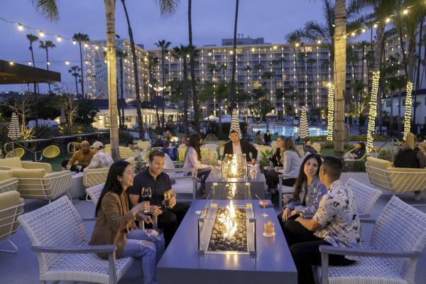 People are seated around fire pits, enjoying conversation and drinks in an outdoor lounge area with string lights, palm trees, and a hotel in the background.