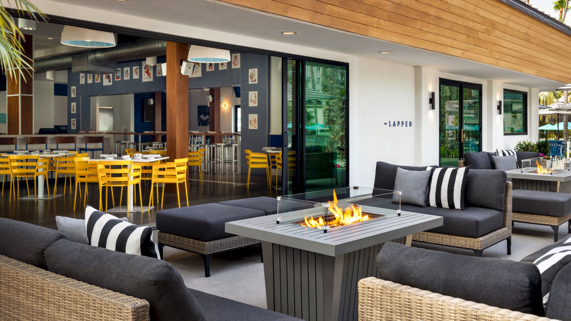 The image shows a modern patio with comfortable seating around a fire pit, and an indoor dining area with yellow chairs.
