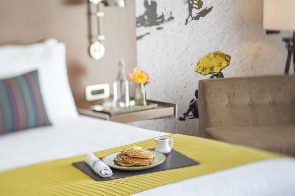 A cozy hotel room with a bed, a side table with flowers and water, and a plate of pancakes served on the bed, ending the sentence.