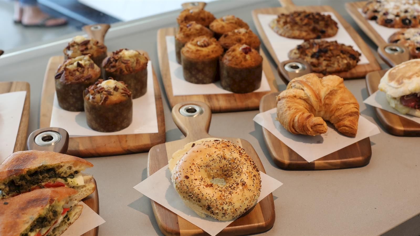 The image shows various baked goods including muffins, croissants, cookies, bagels, and sandwiches arranged on wooden boards on a countertop.