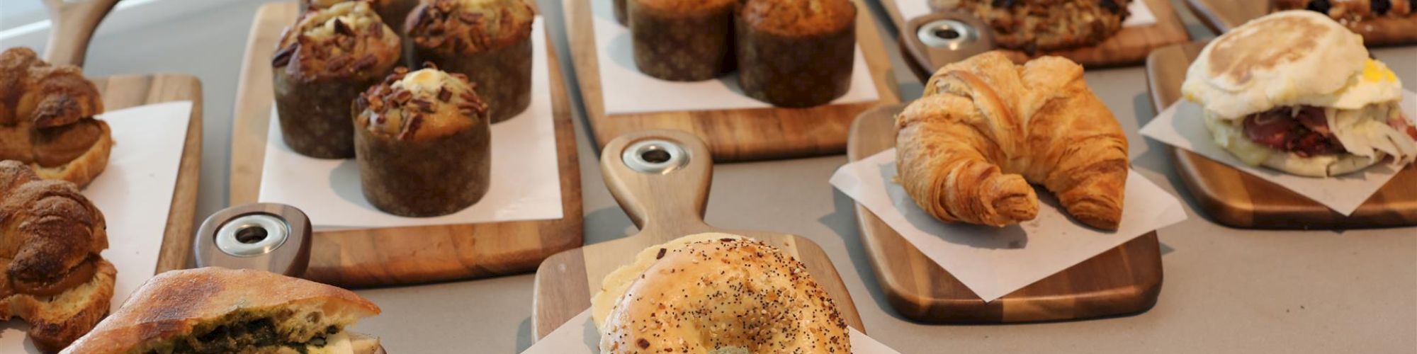 The image shows various baked goods including muffins, croissants, cookies, bagels, and sandwiches arranged on wooden boards on a countertop.