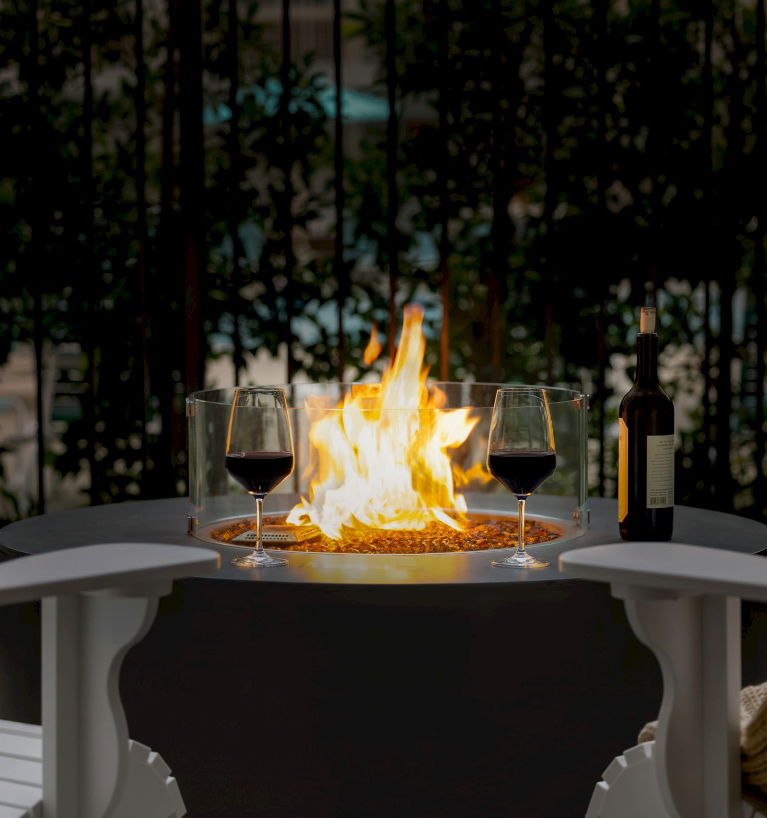 A cozy outdoor fireplace with a glowing fire, two wine glasses on the mantle, and hint of seating and greenery in the background, warm ambiance.