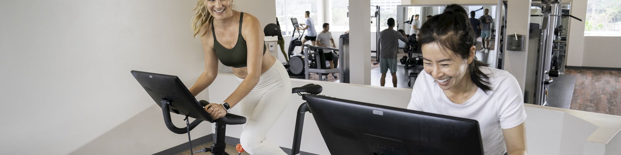 Two women are smiling and riding stationary bikes with screens in a bright gym, filled with other people working out in the background.