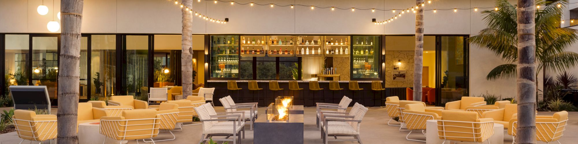 An outdoor lounge area with modern seating, string lights, palm trees, a fire pit in the center, and a well-stocked bar in the background.