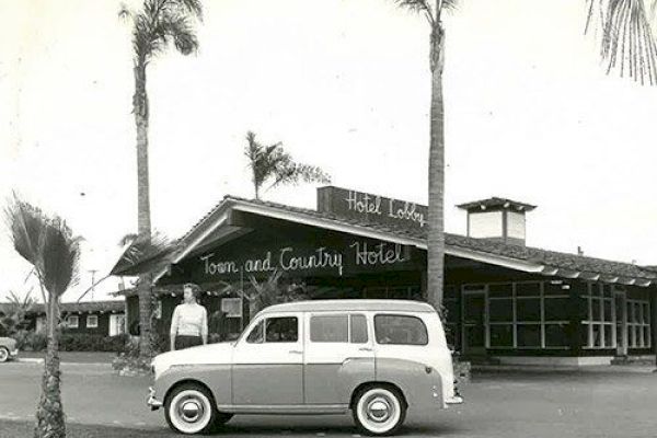 Black and white photo of a vintage car parked in front of the Town and Country Hotel, with a person standing nearby and palm trees around.