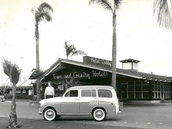 Black and white photo of a vintage car parked in front of the Town and Country Hotel, with a person standing nearby and palm trees around.