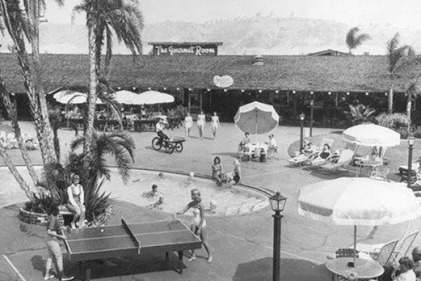An outdoor scene with people playing table tennis, lounging under umbrellas, and swimming, near a building labeled "The Gourmet Room."