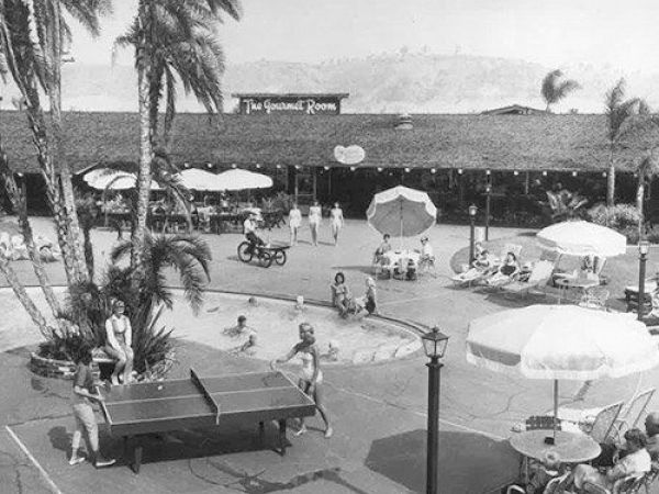 An outdoor scene with people playing table tennis, lounging under umbrellas, and swimming, near a building labeled "The Gourmet Room."