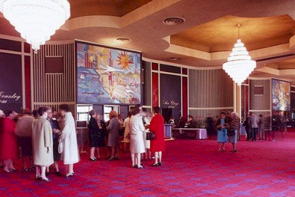 The image depicts a luxurious and spacious indoor setting with people gathered, ornate chandeliers, and vibrant red carpeting.