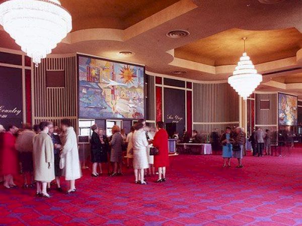 The image depicts a luxurious and spacious indoor setting with people gathered, ornate chandeliers, and vibrant red carpeting.