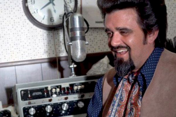 A man with a thick hairstyle and beard smiles while sitting in front of a microphone and vintage audio equipment, with a clock in the background.