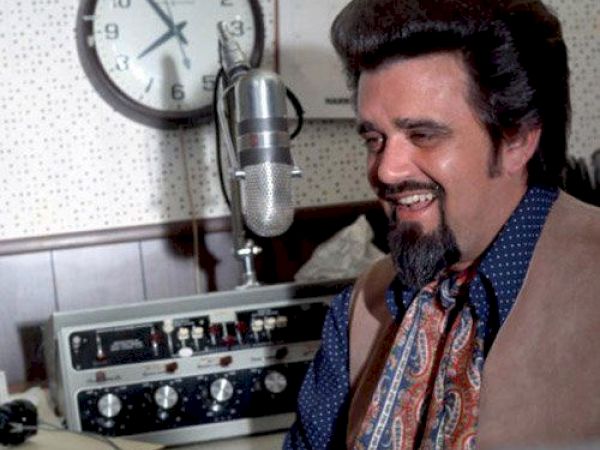 A man with a thick hairstyle and beard smiles while sitting in front of a microphone and vintage audio equipment, with a clock in the background.