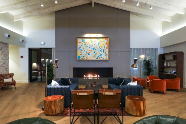 A modern living room with a high ceiling, featuring a central fireplace, abstract artwork, blue sofas, orange chairs, and wooden stools, warmly lit.