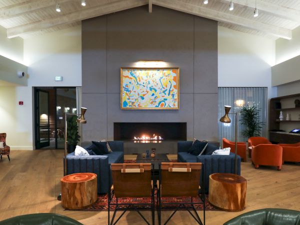 A modern living room with a high ceiling, featuring a central fireplace, abstract artwork, blue sofas, orange chairs, and wooden stools, warmly lit.