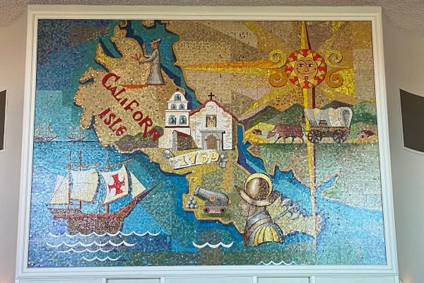This image shows a colorful mural depicting historical scenes and landmarks of California, including a ship, a mission, a road, a miner, and a sun icon.