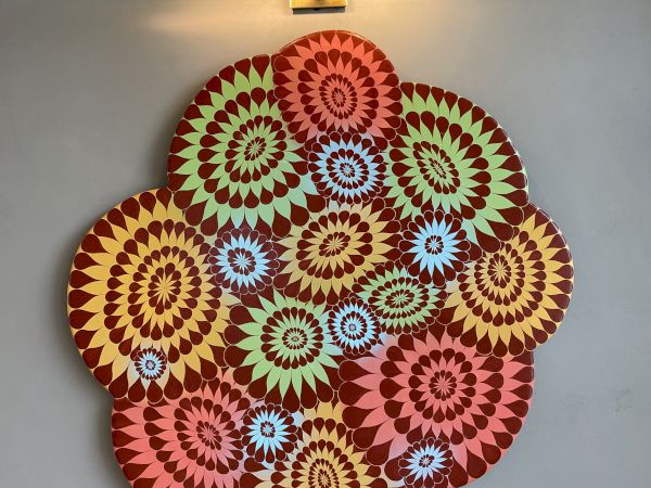 A vibrant, multi-colored geometric artwork is hanging on a wall under a light. The design features intricate spiral patterns in various colors.