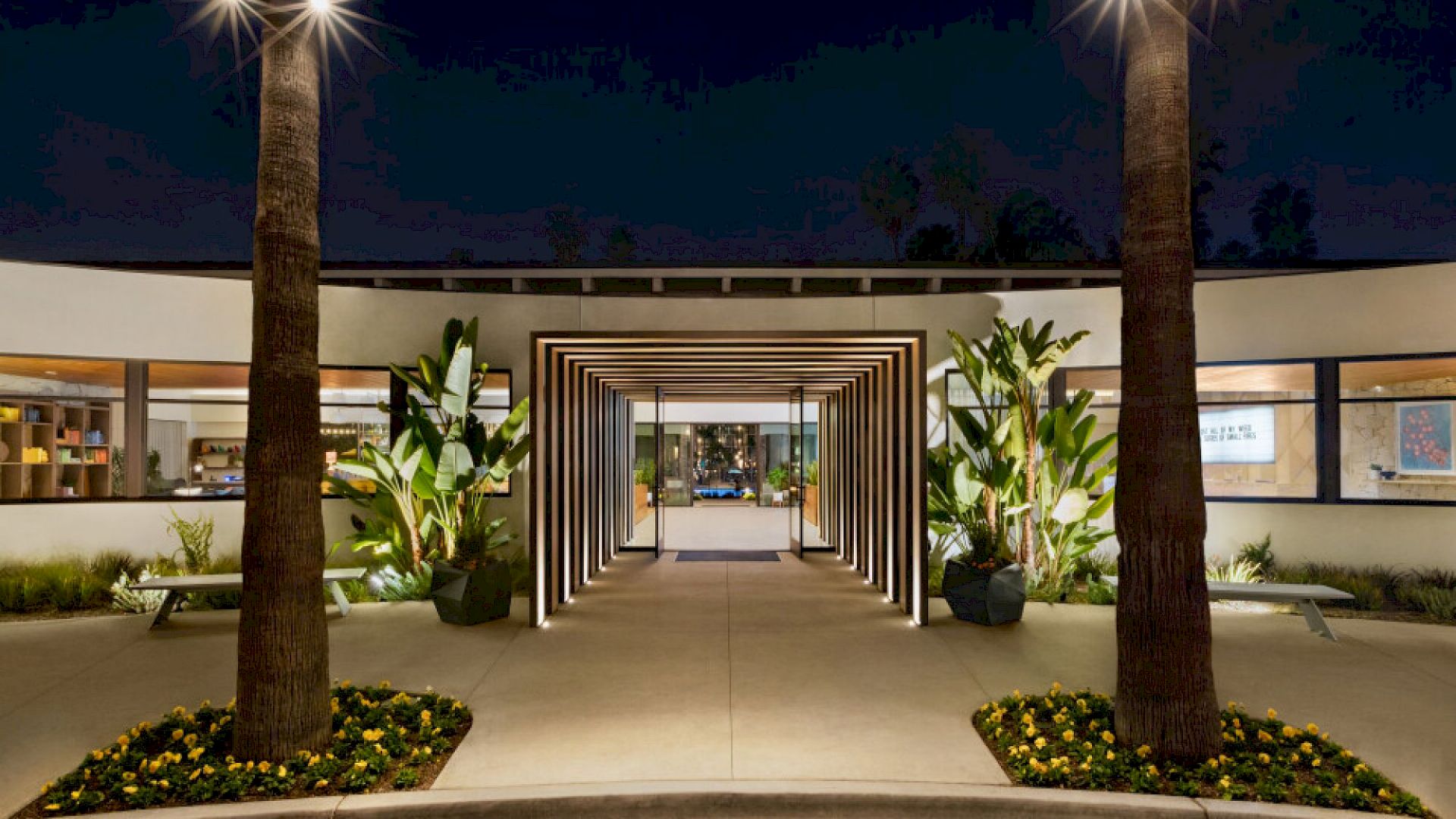 A modern entrance framed by two tall palm trees, illuminated at night, with a pathway lined with greenery and decorative lights.