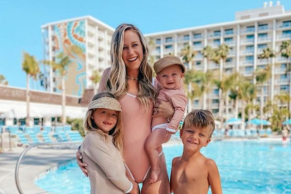 A woman and three children are posing by a pool, smiling. They are all in swimwear, with a hotel building in the background.