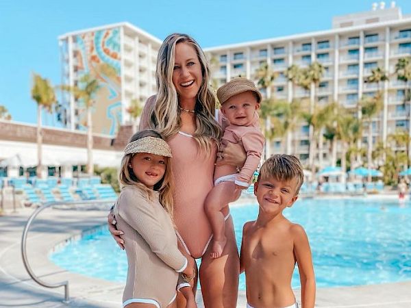 A woman and three children are posing by a pool, smiling. They are all in swimwear, with a hotel building in the background.