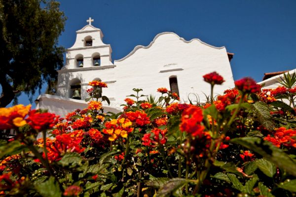 White building with bell tower and cross in background, vibrant orange and red flowers in foreground, clear blue sky.