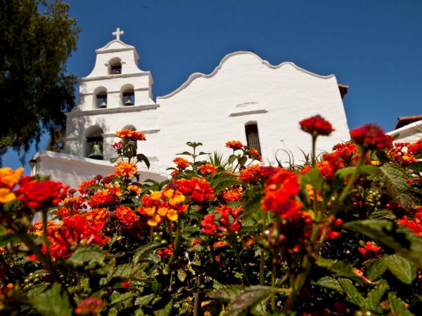 White building with bell tower and cross in background, vibrant orange and red flowers in foreground, clear blue sky.